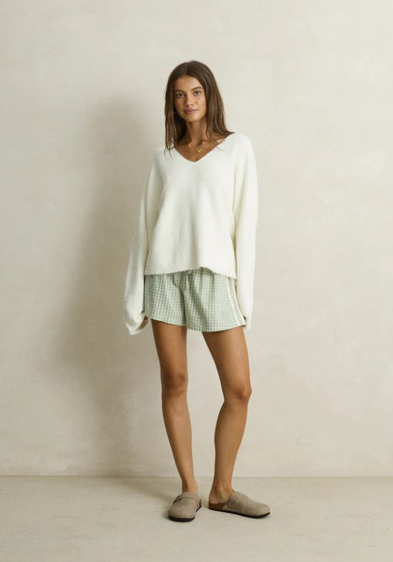 A woman modeling a cozy white knit V-neck sweater paired with green and white gingham shorts, styled with taupe slip-on slippers against a neutral beige wall. Rhythm. Cream. Ladies. Knitwear.