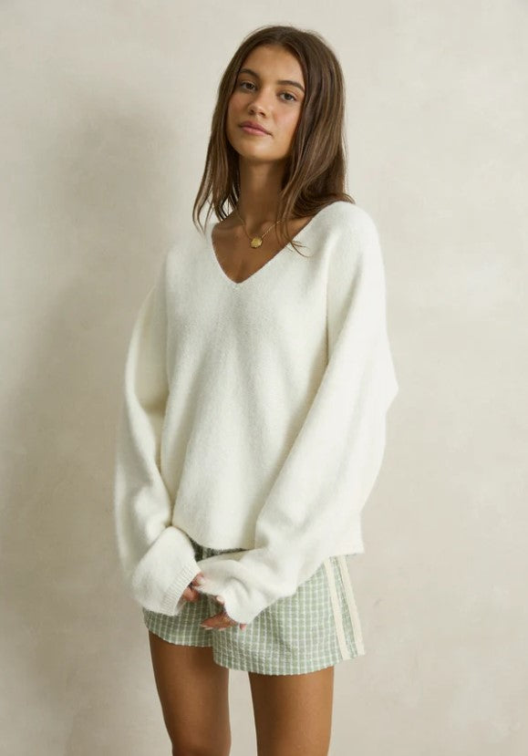 A woman modeling a cozy white knit V-neck sweater paired with green and white gingham shorts, styled with taupe slip-on slippers against a neutral beige wall. Rhythm. Cream. Ladies. Knitwear.