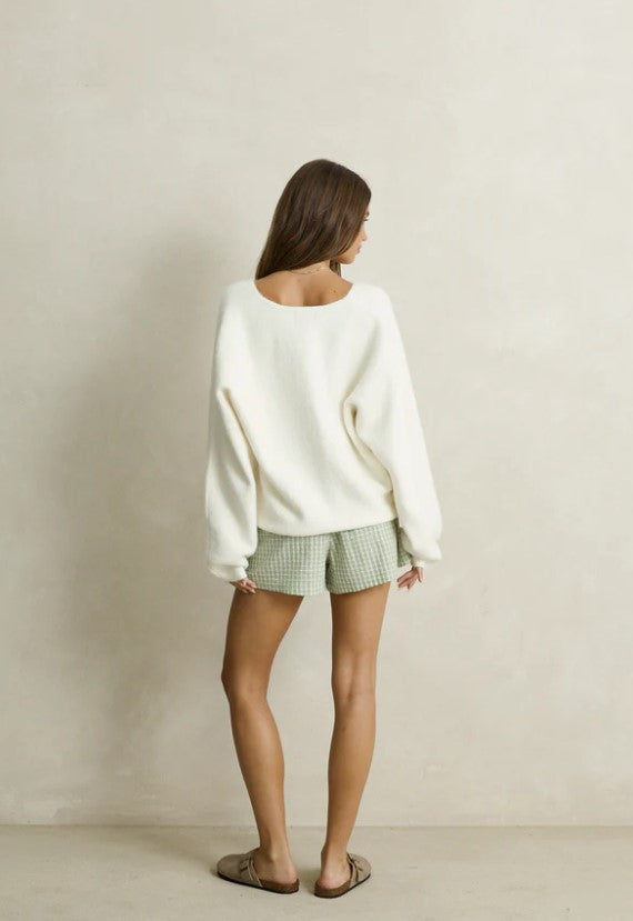 A woman modeling a cozy white knit V-neck sweater paired with green and white gingham shorts, styled with taupe slip-on slippers against a neutral beige wall. Rhythm. Cream. Ladies. Knitwear.