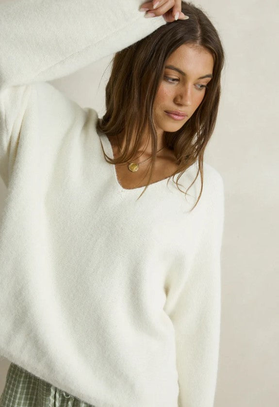 A woman modeling a cozy white knit V-neck sweater paired with green and white gingham shorts, styled with taupe slip-on slippers against a neutral beige wall. Rhythm. Cream. Ladies. Knitwear.