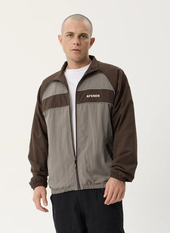 A man wearing a brown and gray color-blocked zip-up jacket with the brand name 'AFENDS' printed in white on the chest, over a white t-shirt and black pants, against a plain light background. SPICY SPORTS SPRAY JACKET OLIVE UNISEX REFLECTIVE PIPING POCKETS