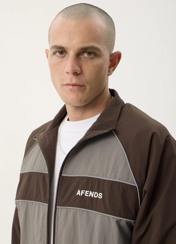 A man wearing a brown and gray color-blocked zip-up jacket with the brand name 'AFENDS' printed in white on the chest, over a white t-shirt and black pants, against a plain light background. SPICY SPORTS SPRAY JACKET OLIVE UNISEX REFLECTIVE PIPING POCKETS