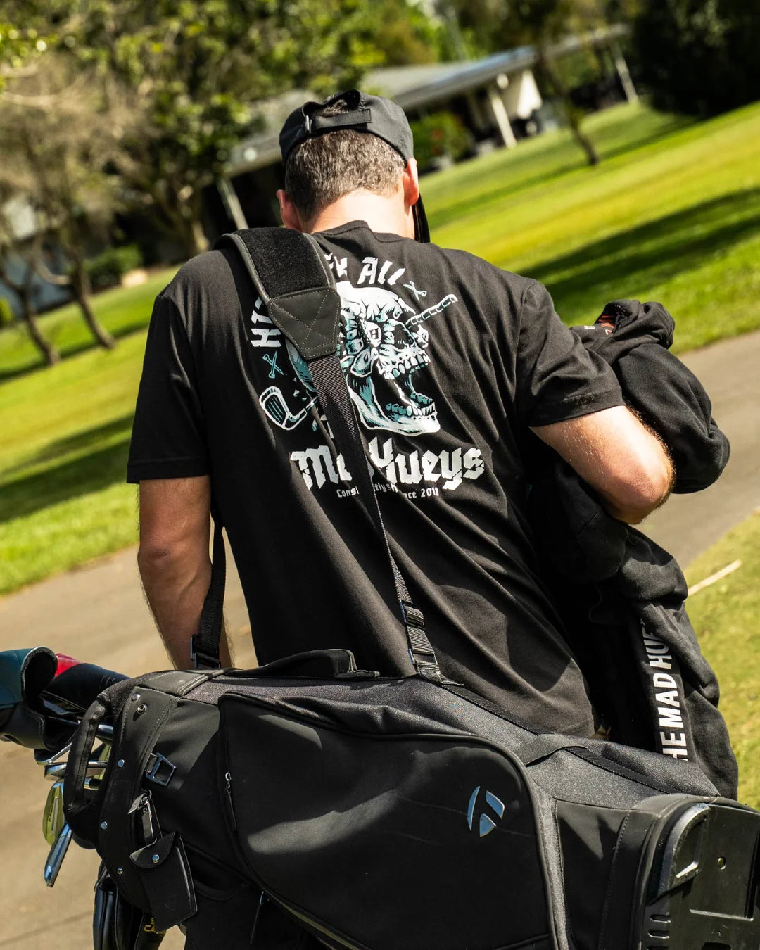 A man seen from behind wearing a black graphic t-shirt and a black baseball cap, carrying a large black golf bag on his shoulder in a sunny park setting.