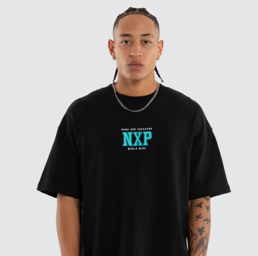 A person shown from the back wearing a black oversized T-shirt with a large turquoise and white graphic reading 'NENA AND PASADENA Upscale Supply WORLD WIDE MISSION' on a light gray studio background. WEST LOOPBACK RAPTOR TEE ANTHRACITE