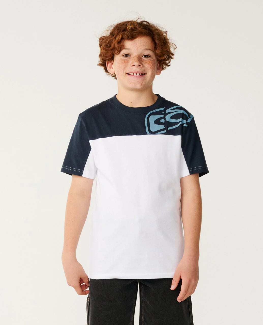 A young boy with curly red hair stands against a light off-white background wearing a colorblock t-shirt. The shirt features a navy blue upper block and sleeves with a light blue graphic logo on the right chest, and a solid white lower block.