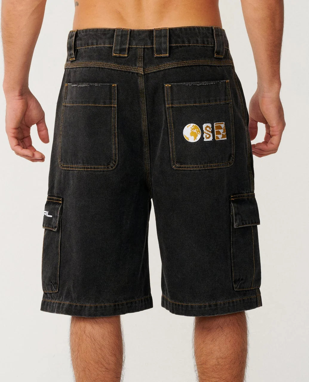Rear view of a person wearing dark washed cargo shorts with a circular gold, white, and blue patch on the back pocket, photographed against a light gray studio background.