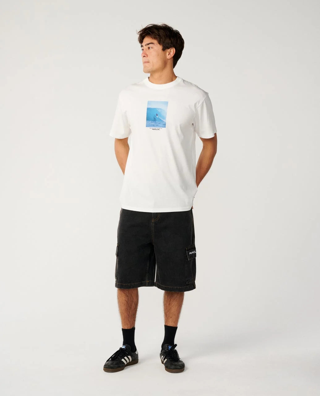 A man modeling a white crewneck t-shirt with a blue graphic print on the chest, paired with black cargo shorts, black socks, and black sneakers, against a plain white studio background.