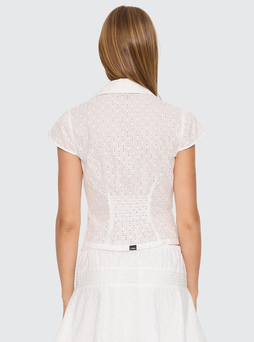 A woman modeling a white eyelet two-piece outfit consisting of a short-sleeve collared crop top with a front zipper and a matching mini skirt with a drawstring, against a light gray studio background. DANICA BRODERIE ANGLAISE TOP THRILLS LADIES TOP T-SHIRT WHITE COLLAR ZIP