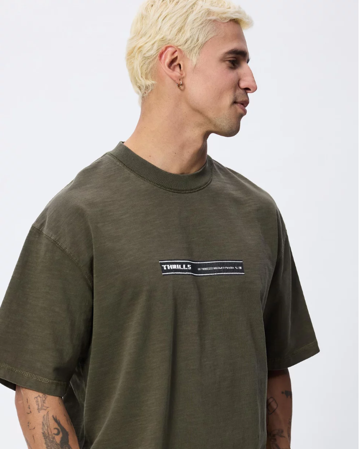 A man with platinum blonde hair wearing an oversized olive green t-shirt with a 'THRILLER' graphic on the chest, paired with dark brown pants, photographed against a light gray studio background. THRILLS PARADISE BUILT OVERSIZE BOX CROP TEE MENS OLIVE NIGHT KHAKI GREEN