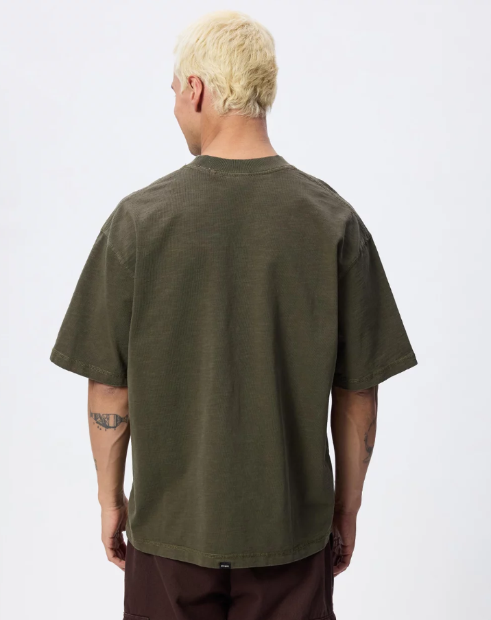 A man with platinum blonde hair wearing an oversized olive green t-shirt with a 'THRILLER' graphic on the chest, paired with dark brown pants, photographed against a light gray studio background. THRILLS PARADISE BUILT OVERSIZE BOX CROP TEE MENS OLIVE NIGHT KHAKI GREEN