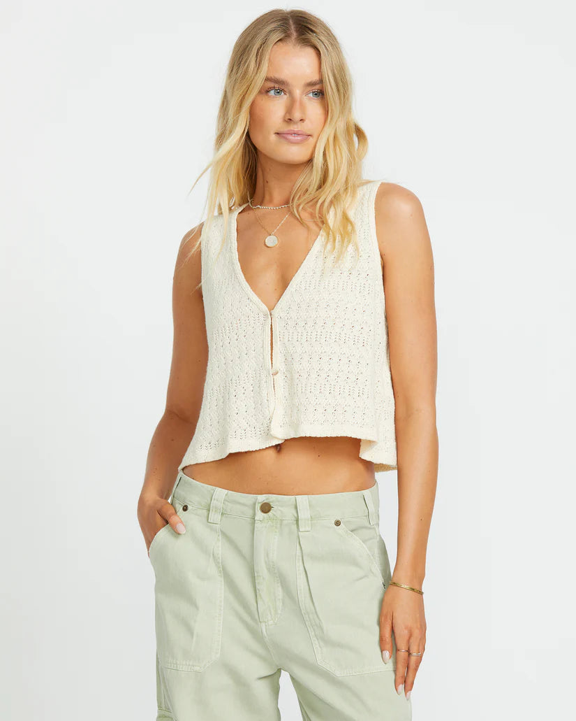 Woman wearing a white sleeveless knitted top and light green pants on a plain background