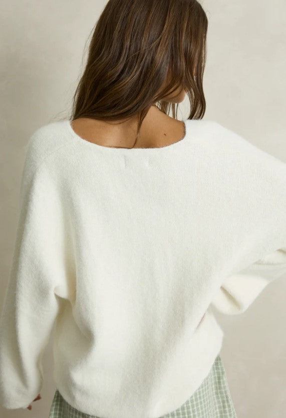 A woman modeling a cozy white knit V-neck sweater paired with green and white gingham shorts, styled with taupe slip-on slippers against a neutral beige wall. Rhythm. Cream. Ladies. Knitwear.