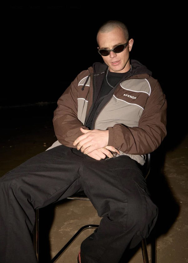A man wearing a brown and gray color-blocked zip-up jacket with the brand name 'AFENDS' printed in white on the chest, over a white t-shirt and black pants, against a plain light background. SPICY SPORTS SPRAY JACKET OLIVE UNISEX REFLECTIVE PIPING POCKETS