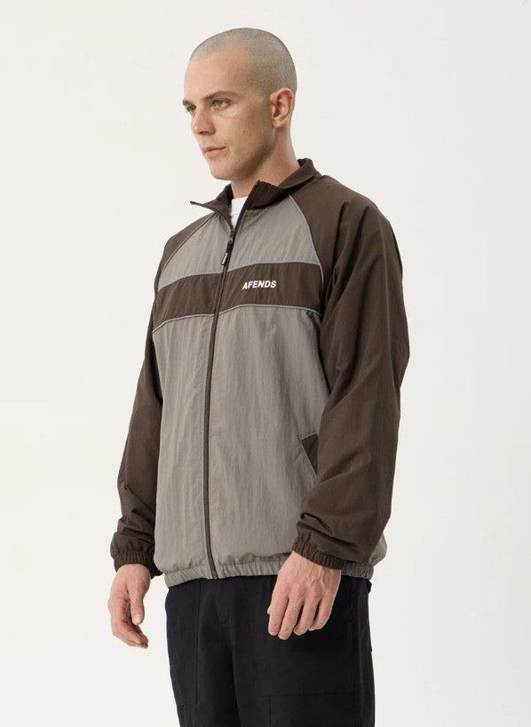A man wearing a brown and gray color-blocked zip-up jacket with the brand name 'AFENDS' printed in white on the chest, over a white t-shirt and black pants, against a plain light background. SPICY SPORTS SPRAY JACKET OLIVE UNISEX REFLECTIVE PIPING POCKETS