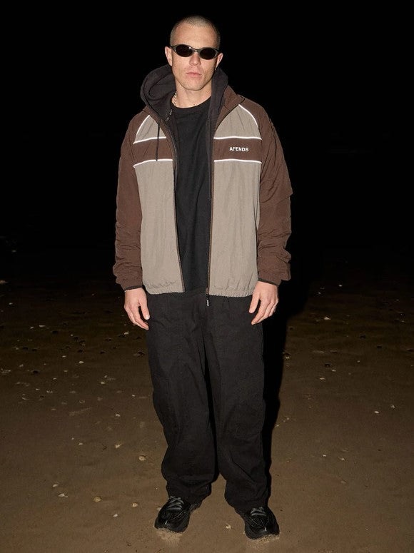 A man wearing a brown and gray color-blocked zip-up jacket with the brand name 'AFENDS' printed in white on the chest, over a white t-shirt and black pants, against a plain light background. SPICY SPORTS SPRAY JACKET OLIVE UNISEX REFLECTIVE PIPING POCKETS