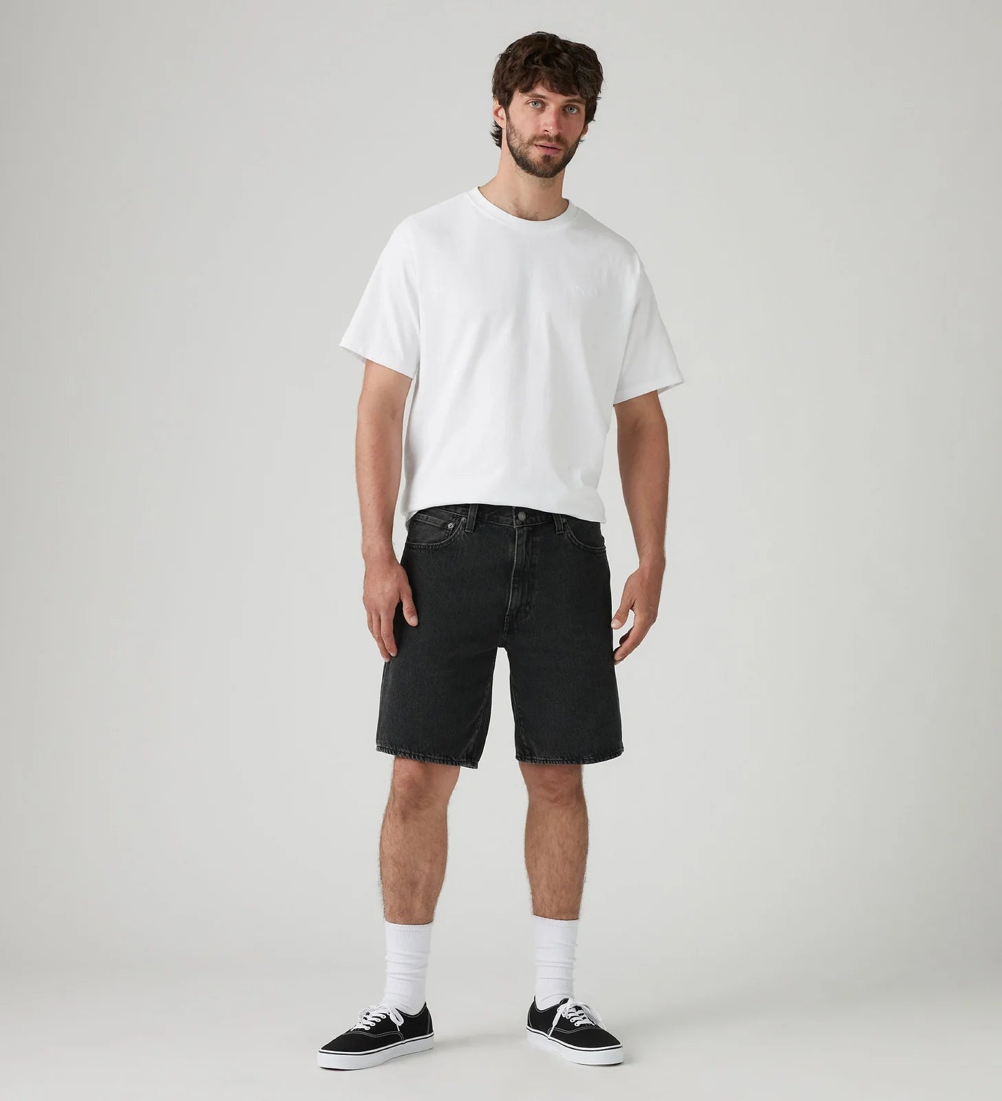 A man modeling a white crewneck t-shirt, black denim shorts, white socks, and black low-top sneakers against a light gray studio background.