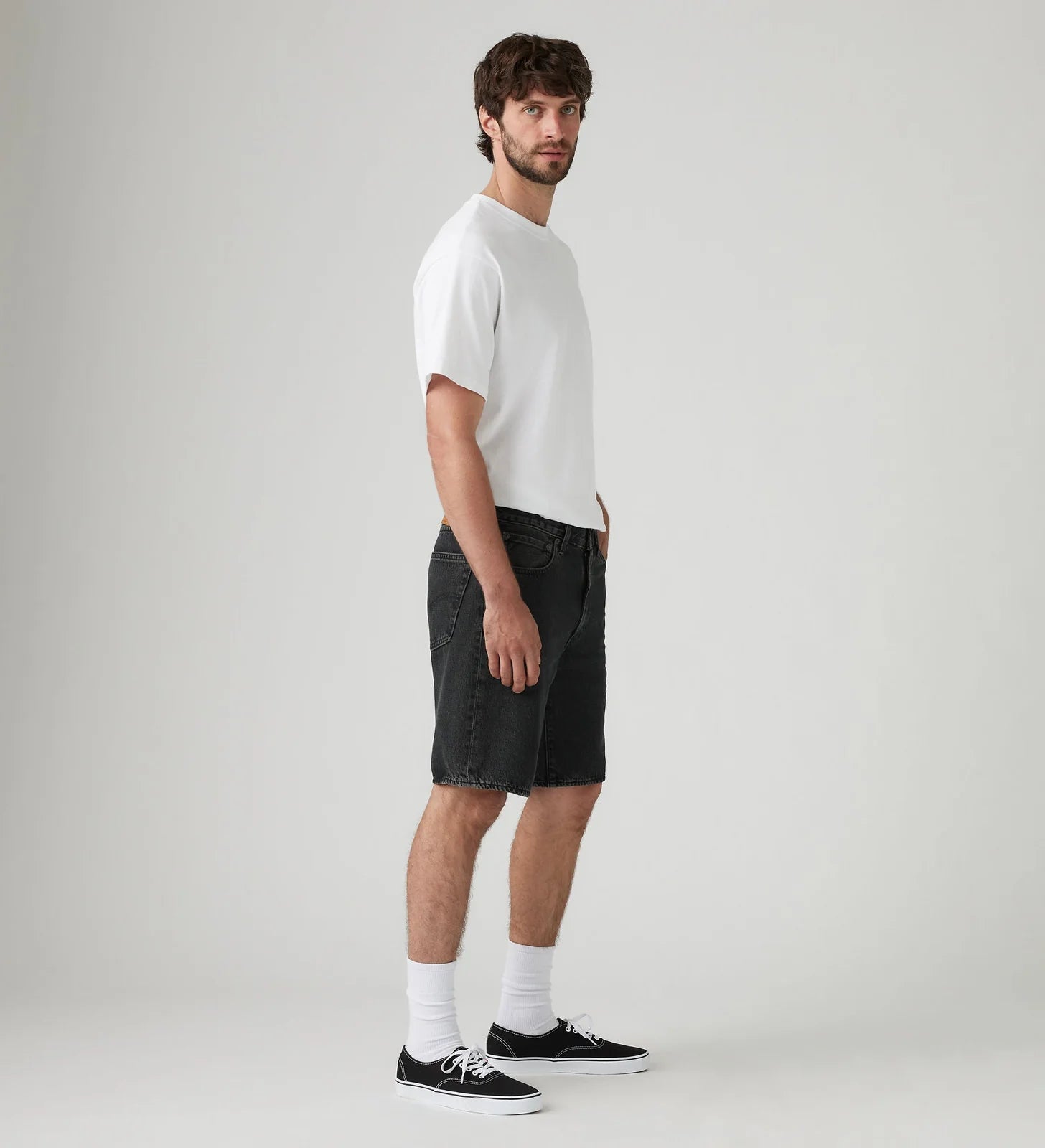 A man stands in profile wearing a plain white short-sleeve T-shirt, dark charcoal denim shorts, white socks, and black low-top sneakers against a light gray studio background.