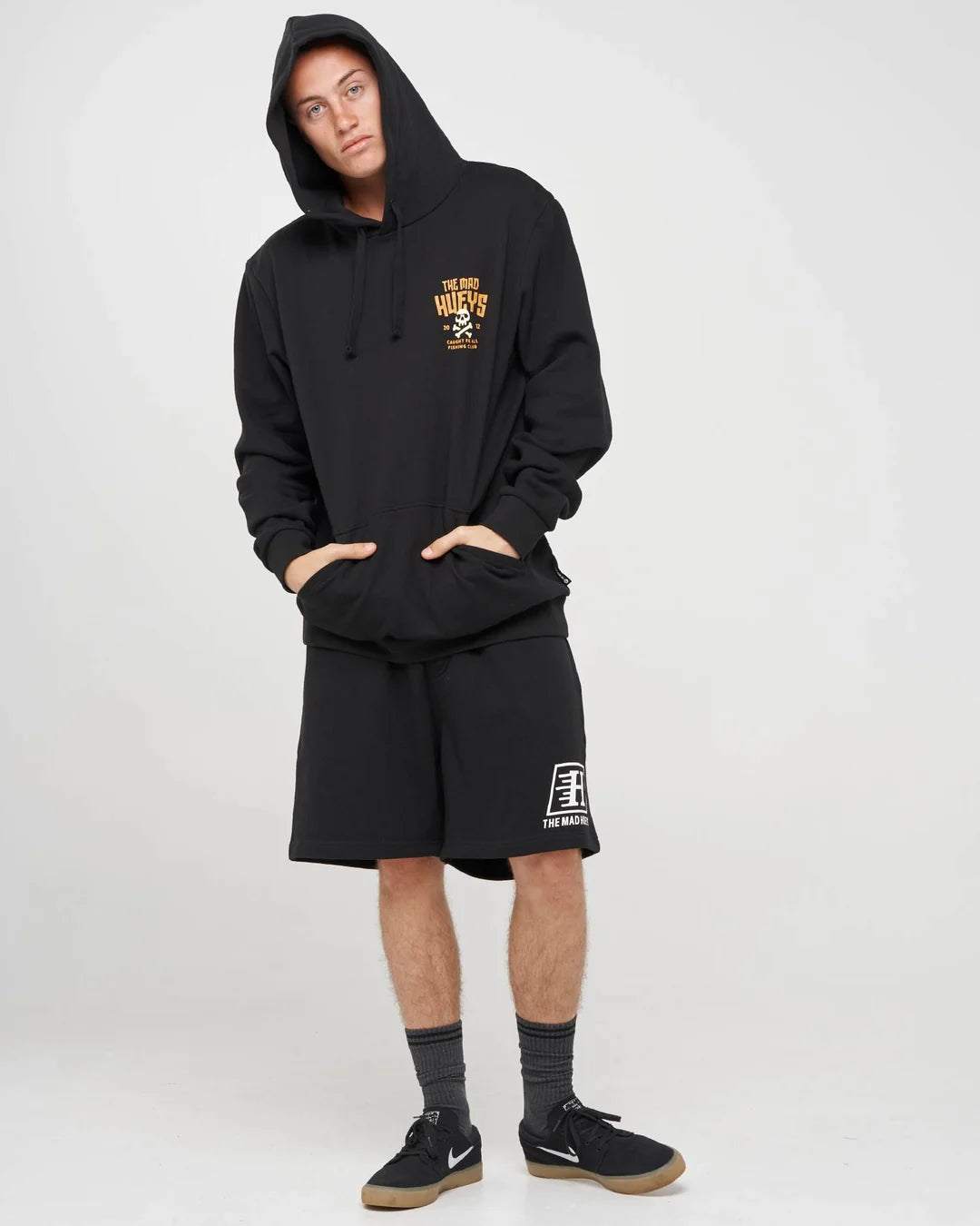 A model wearing a black pullover hoodie with a yellow and white graphic on the left chest, matching black shorts with a white graphic on the left leg, black crew socks, and black sneakers, standing against a light gray studio background.