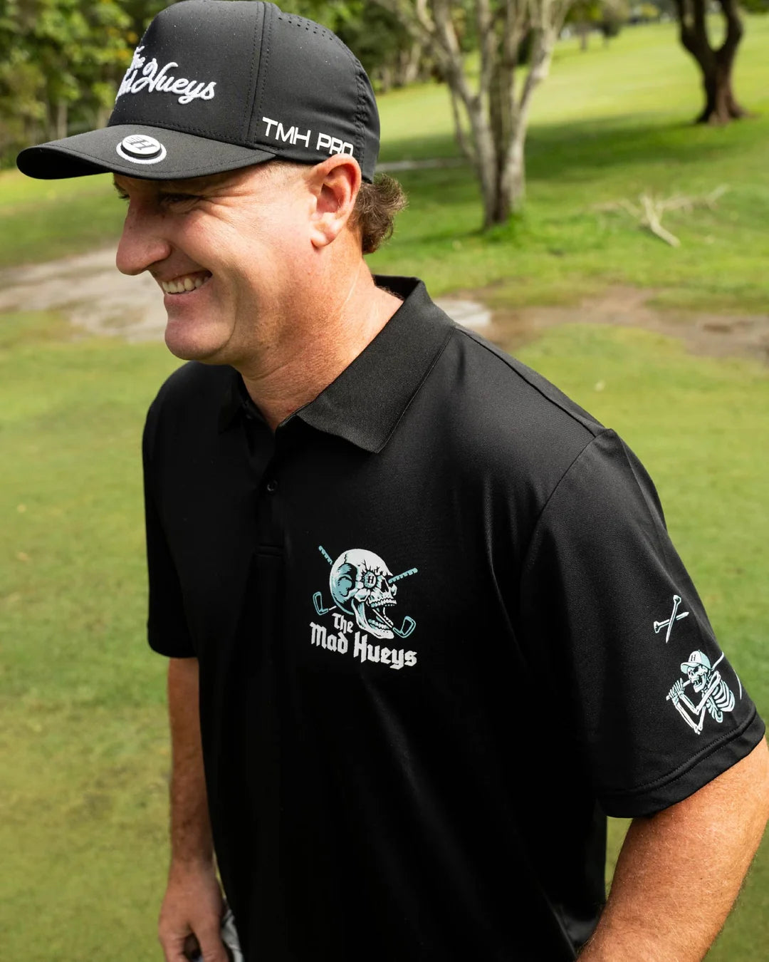 A man on a golf course wearing a black polo shirt and matching black cap with white and teal graphics. The polo shirt features a 'Mad Hueys' skull logo on the left chest, and the cap has 'Mad Hueys' and 'TMH PRO' branding.
