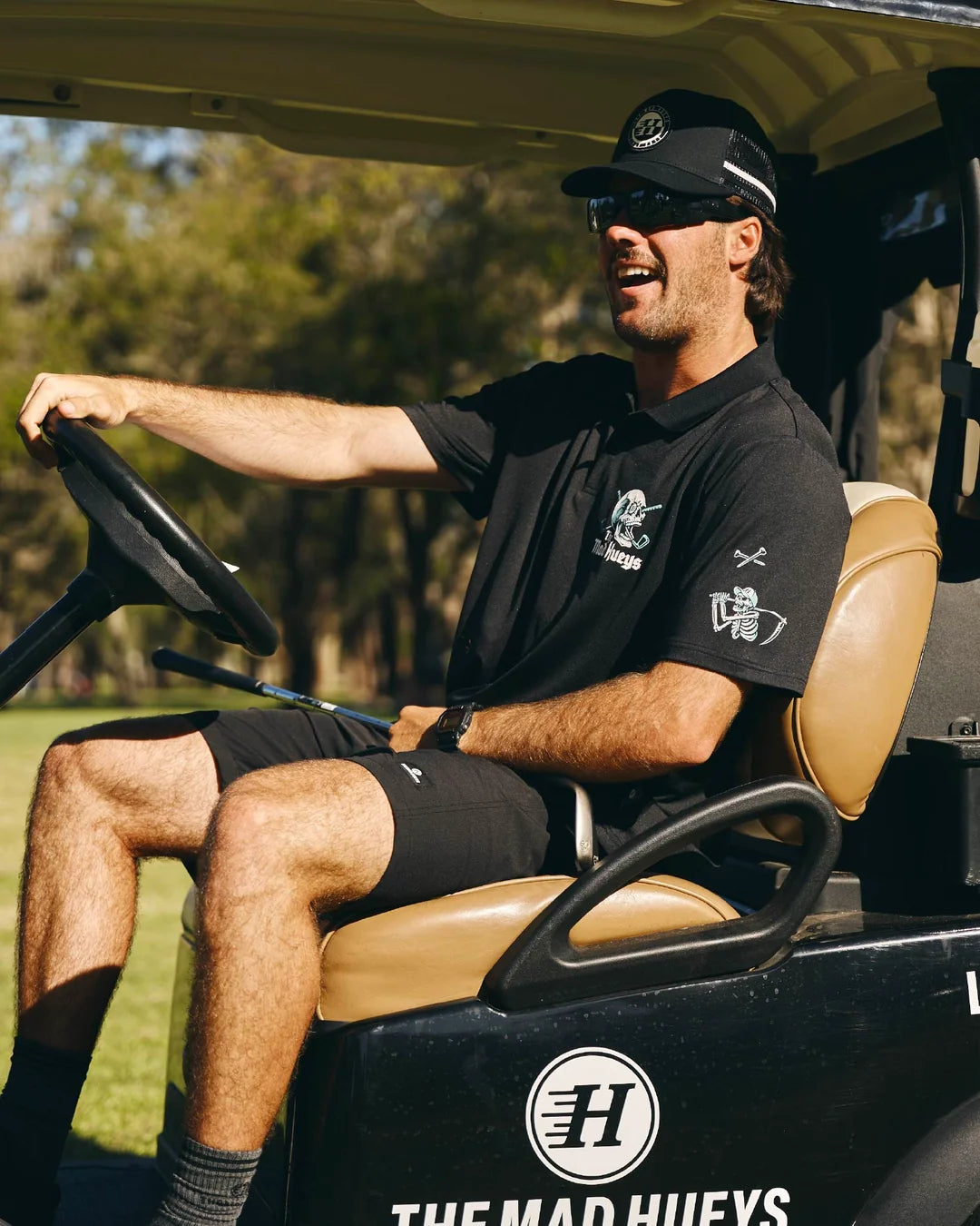 A man wearing a black polo shirt, black shorts, and a black cap sits in a golf cart on a golf course. The shirt features white and silver graphics, and the cart has 'THE MAD HUEYS' branding visible on the side.