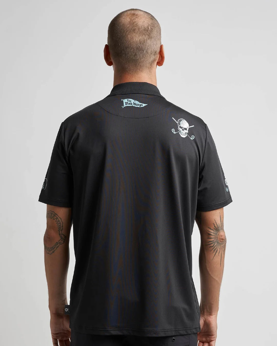 A man viewed from behind wearing a black short-sleeve polo shirt with white graphics, including a skull and crossed golf clubs on the left shoulder and a logo on the back collar.