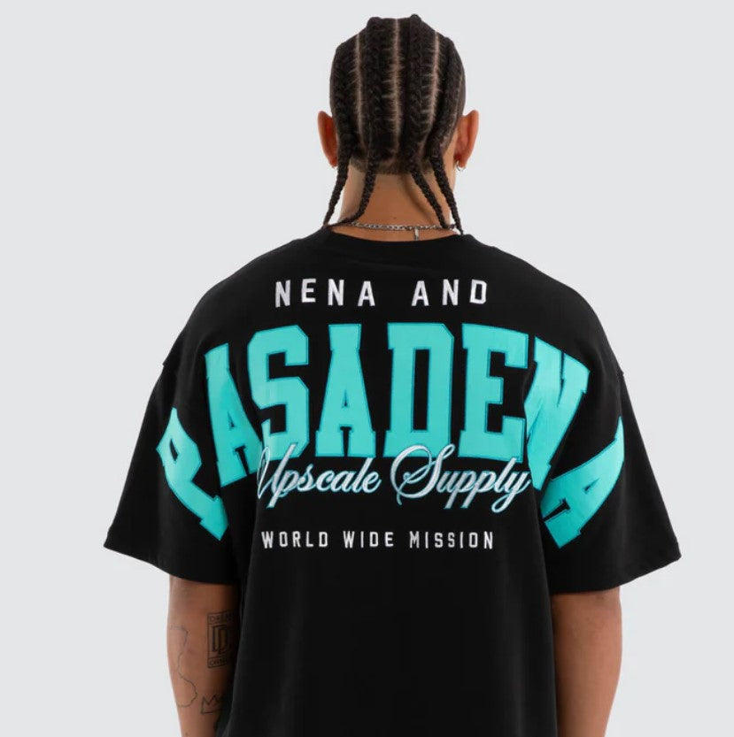 A person shown from the back wearing a black oversized T-shirt with a large turquoise and white graphic reading 'NENA AND PASADENA Upscale Supply WORLD WIDE MISSION' on a light gray studio background. WEST LOOPBACK RAPTOR TEE ANTHRACITE