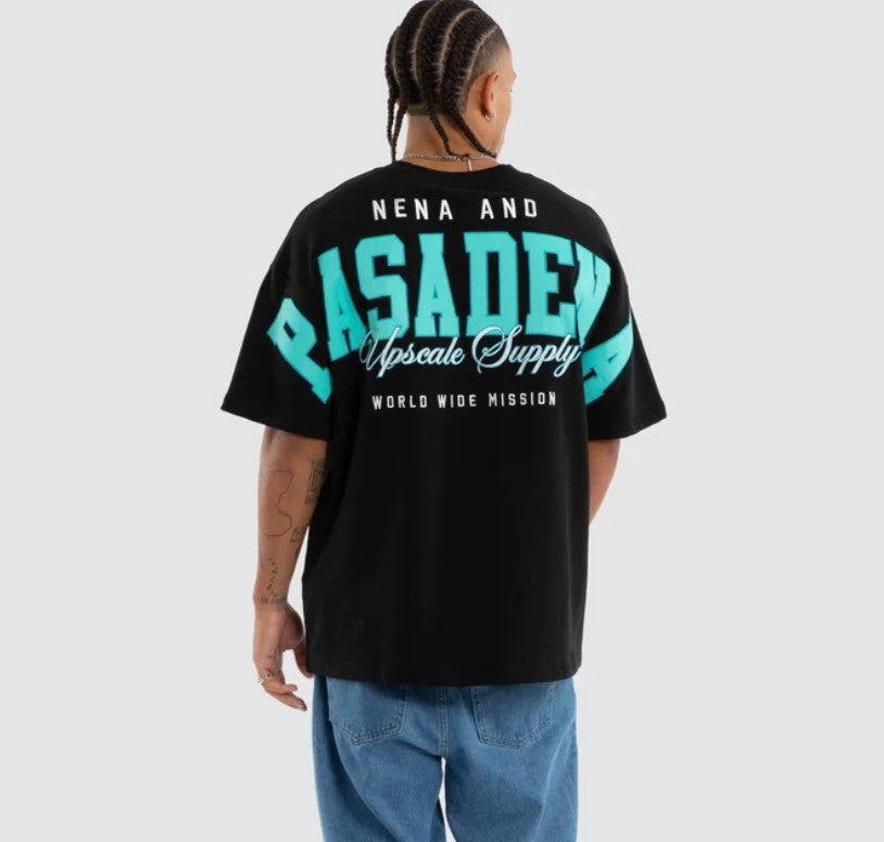 A person shown from the back wearing a black oversized T-shirt with a large turquoise and white graphic reading 'NENA AND PASADENA Upscale Supply WORLD WIDE MISSION' on a light gray studio background. WEST LOOPBACK RAPTOR TEE ANTHRACITE