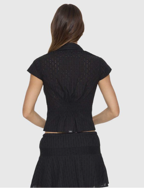 A woman modeling a black ribbed short-sleeve crop top with a front zipper, paired with a matching black textured skirt, against a plain white background. DANICA BRODERIE ANGLAISE TOP THRILLS BLACK LADIES TOP COLLAR ZIP