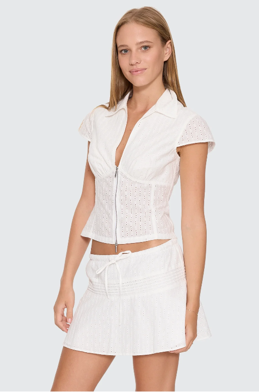 A woman modeling a white eyelet two-piece outfit consisting of a short-sleeve collared crop top with a front zipper and a matching mini skirt with a drawstring, against a light gray studio background. DANICA BRODERIE ANGLAISE TOP THRILLS LADIES TOP T-SHIRT WHITE COLLAR ZIP