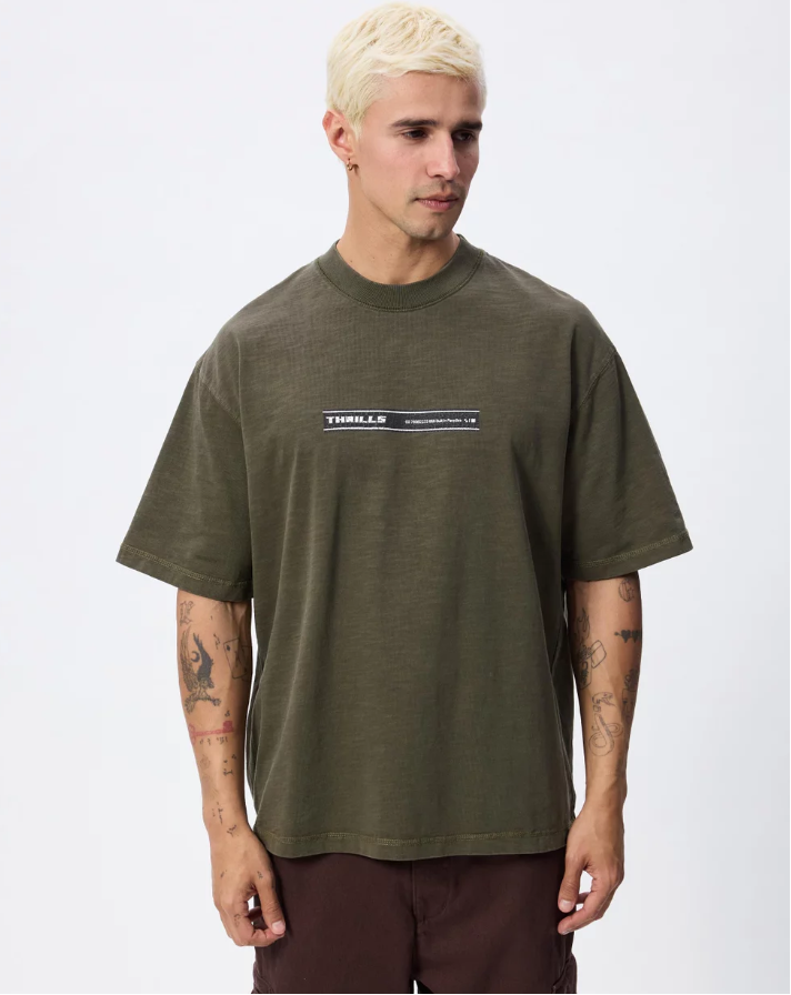 A man with platinum blonde hair wearing an oversized olive green t-shirt with a 'THRILLER' graphic on the chest, paired with dark brown pants, photographed against a light gray studio background. THRILLS PARADISE BUILT OVERSIZE BOX CROP TEE MENS OLIVE NIGHT KHAKI GREEN