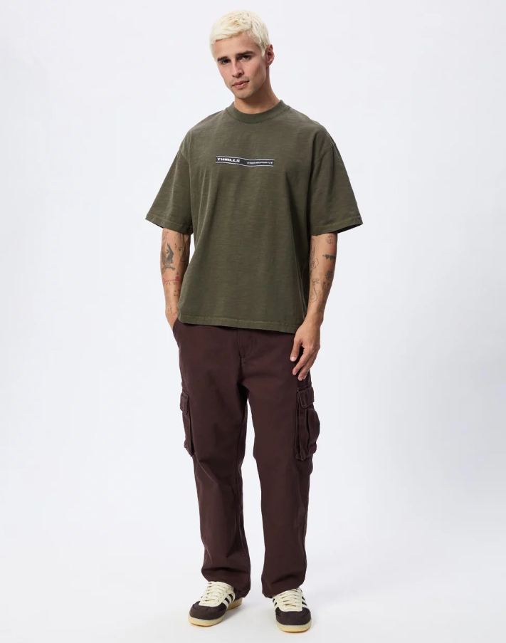 A man with platinum blonde hair wearing an oversized olive green t-shirt with a 'THRILLER' graphic on the chest, paired with dark brown pants, photographed against a light gray studio background. THRILLS PARADISE BUILT OVERSIZE BOX CROP TEE MENS OLIVE NIGHT KHAKI GREEN