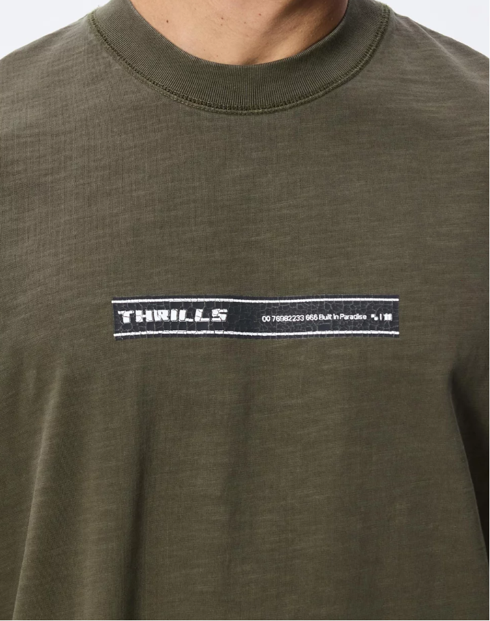 A man with platinum blonde hair wearing an oversized olive green t-shirt with a 'THRILLER' graphic on the chest, paired with dark brown pants, photographed against a light gray studio background. THRILLS PARADISE BUILT OVERSIZE BOX CROP TEE MENS OLIVE NIGHT KHAKI GREEN