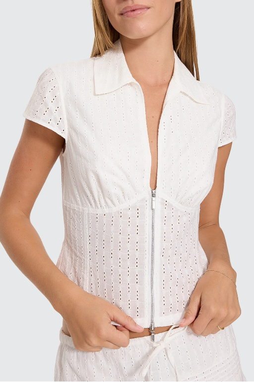A woman modeling a white eyelet two-piece outfit consisting of a short-sleeve collared crop top with a front zipper and a matching mini skirt with a drawstring, against a light gray studio background. DANICA BRODERIE ANGLAISE TOP THRILLS LADIES TOP T-SHIRT WHITE COLLAR ZIP