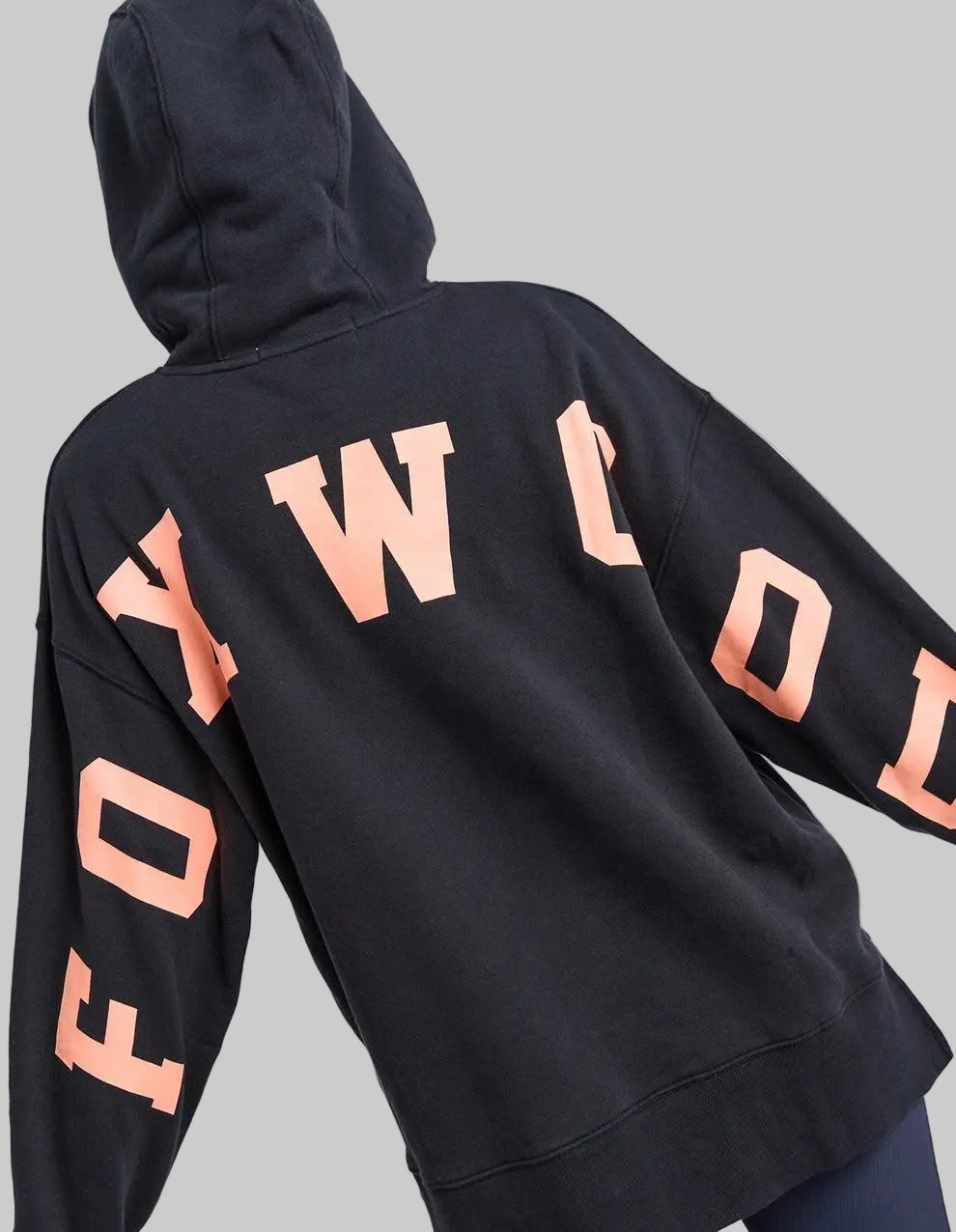 A black pullover hoodie shown from the back, featuring large peach-colored varsity-style letters spelling 'FOXWOOD' across the upper back and sleeves, worn by a model against a light gray studio background.