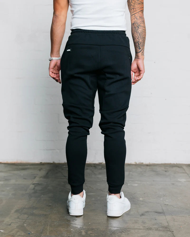 Person wearing wndrr black jogger pants and white sneakers against a plain background